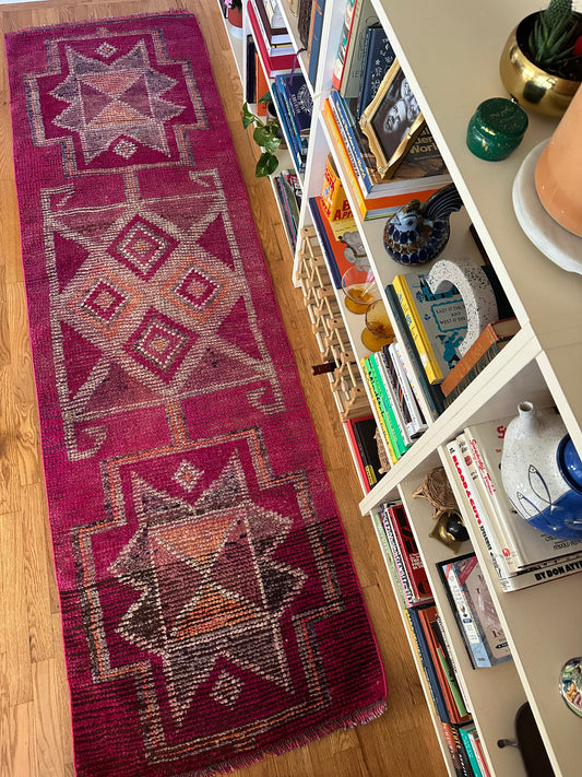 Pink geometric patterned rug on a wooden floor next to a bookshelf with decorative items.